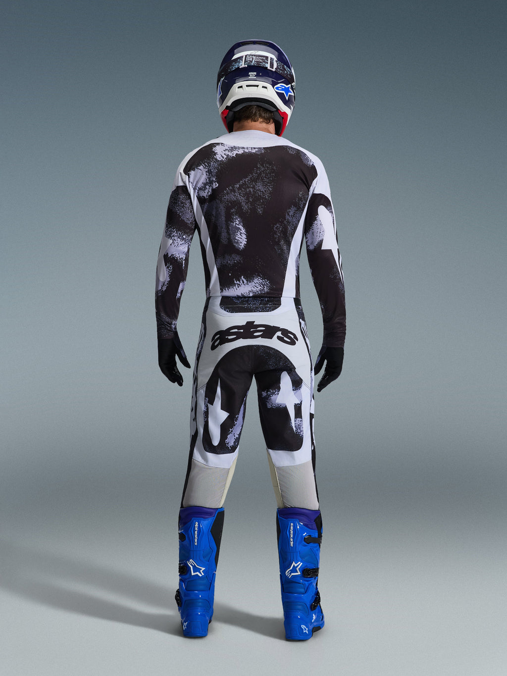 Motocross rider from behind wearing the Alpinestars 2026 Racer Lahnd Jersey and matching pants in Iron Camo, featuring a black and white splatter pattern, paired with bright blue boots and a coordinating helmet, complete gear set for off-road racing