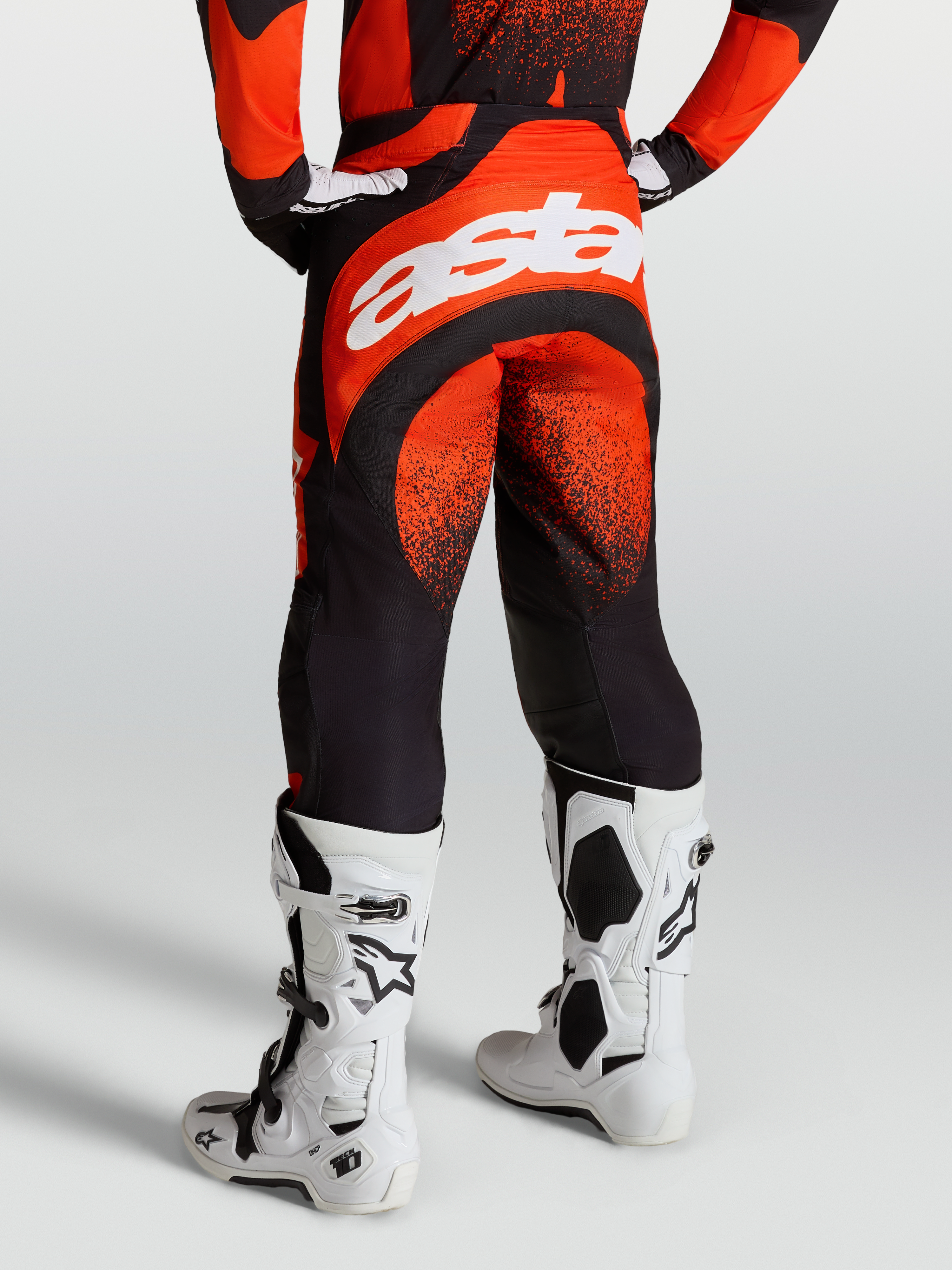 Alpinestars Techstar Swan broek, heren motorcrossbroek, rood en zwart, wit Astar-logo, gedragen met witte Tech 10 laarzen.