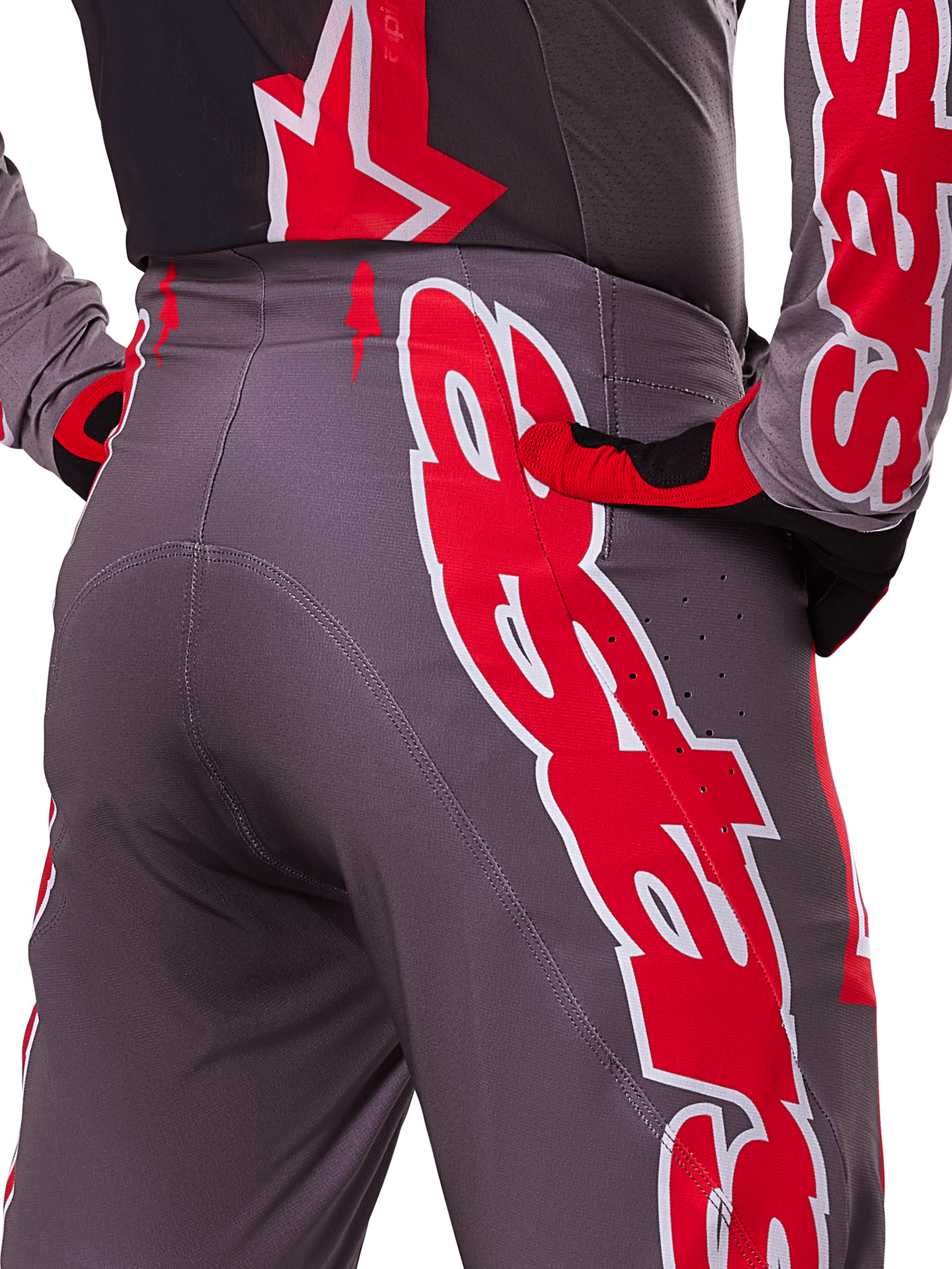 Alpinestars 2025 Supertech Lipan Pants, pantalon de motocross, Smoke Bright Red, vue rapprochée de l'arrière montrant un tissu gris foncé avec des graphismes rouges vifs, porté avec des gants et un maillot rouges et noirs assortis, doté de perforations de ventilation et de coutures ergonomiques pour des performances tout-terrain