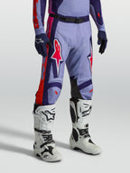Alpinestars Racer Air Hunt Broek, geventileerde paarse en roze motorcrossbroek gedragen door een model met witte motorlaarzen, technische off-road race-uitrusting.