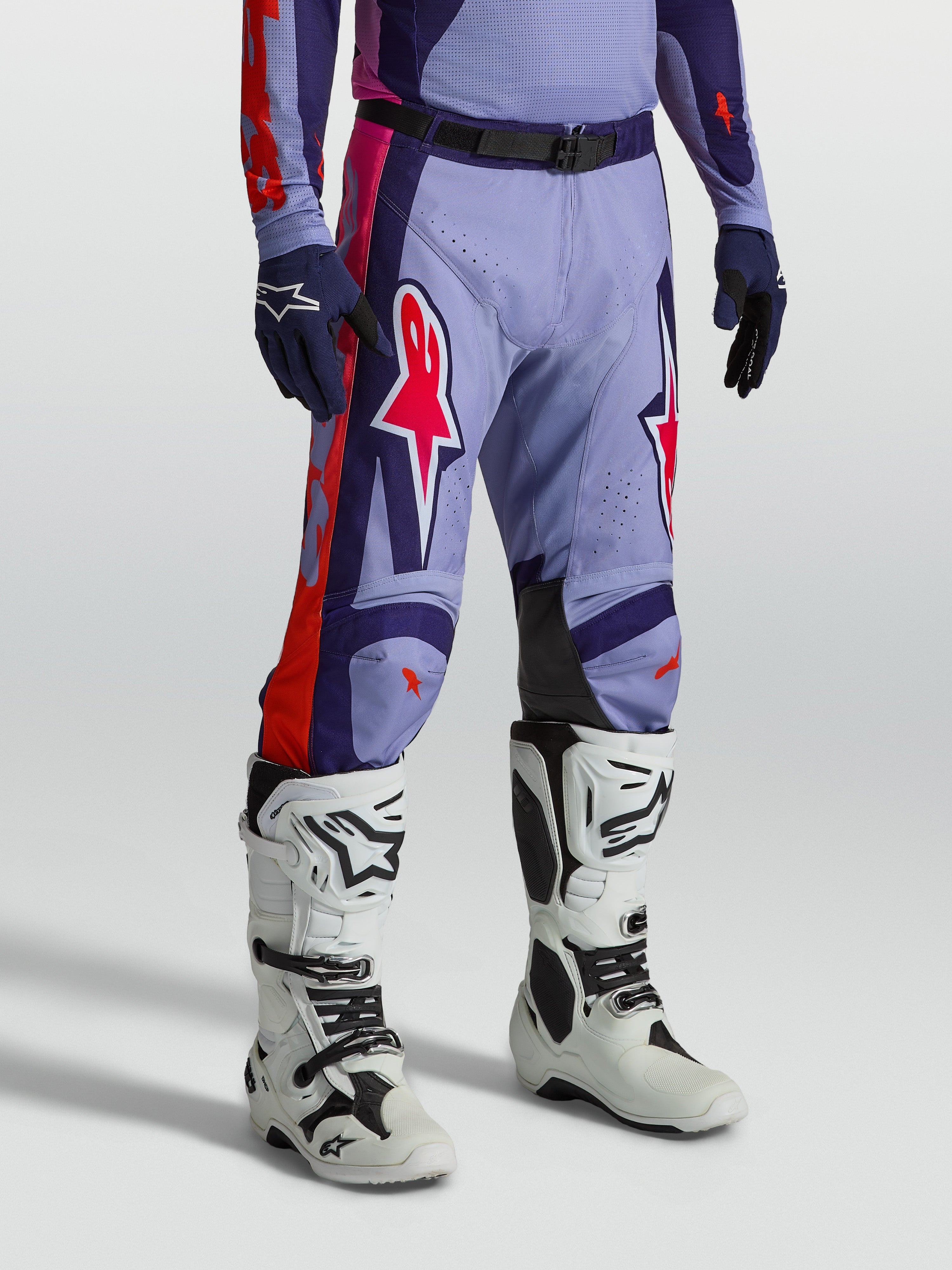 Alpinestars Racer Air Hunt Broek, geventileerde paarse en roze motorcrossbroek gedragen door een model met witte motorlaarzen, technische off-road race-uitrusting.