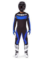 Pilote de motocross portant l'ensemble Alpinestars 2025 Techstar Dreem, maillot noir avec panneau de poitrine et manches bleus, pantalon noir et bleu assorti, casque noir avec lunettes et bottes blanches, tenue de motocross complète coordonnée pour la course tout-terrain.