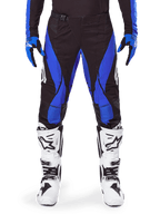 Pantalon Alpinestars Techstar Dreem 2025, pantalon de motocross, noir et bleu, porté avec un maillot, des gants assortis et des bottes blanches, doté de panneaux latéraux bleus et d'une coupe de course athlétique, matériau durable pour la course tout-terrain