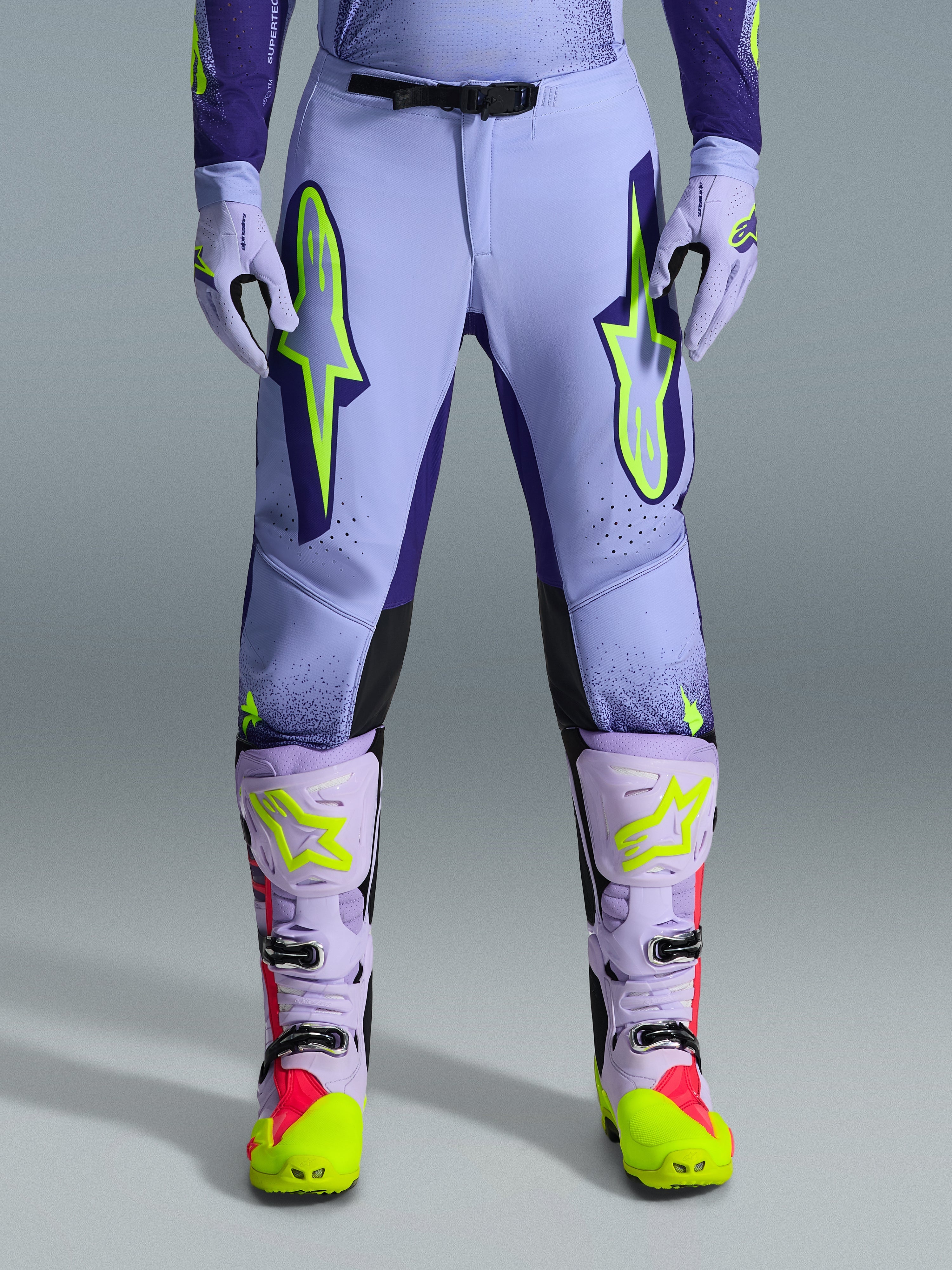 Alpinestars 2026 Supertech Scenz Hose, Motocross-Hose, Lila Gelb Fluo, mit großen neongelben Logos auf den Oberschenkeln, getragen mit passenden lila-gelben Motocross-Stiefeln und -Handschuhen, athletisches Performance-Design für den Offroad-Rennsport