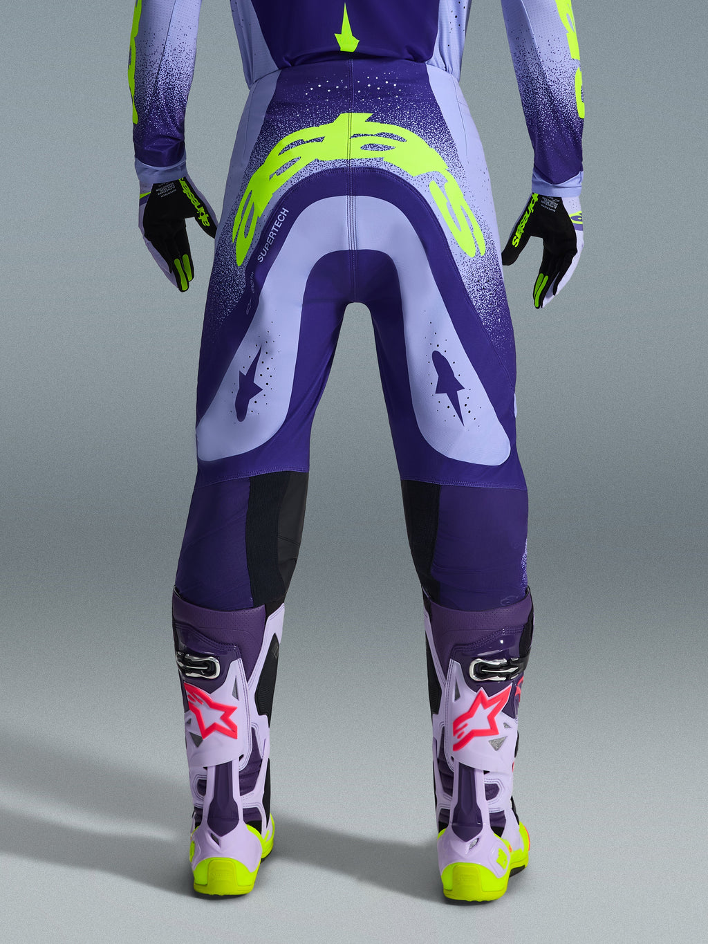 Alpinestars 2026 Supertech Scenz Hose, Motocross-Hose, Lila und Gelb Fluo, Rückansicht mit großem gelben Logo am Gesäß, getragen mit passendem Trikot, Handschuhen und Schutzstiefeln, athletische Passform für professionellen Motocross-Rennsport.