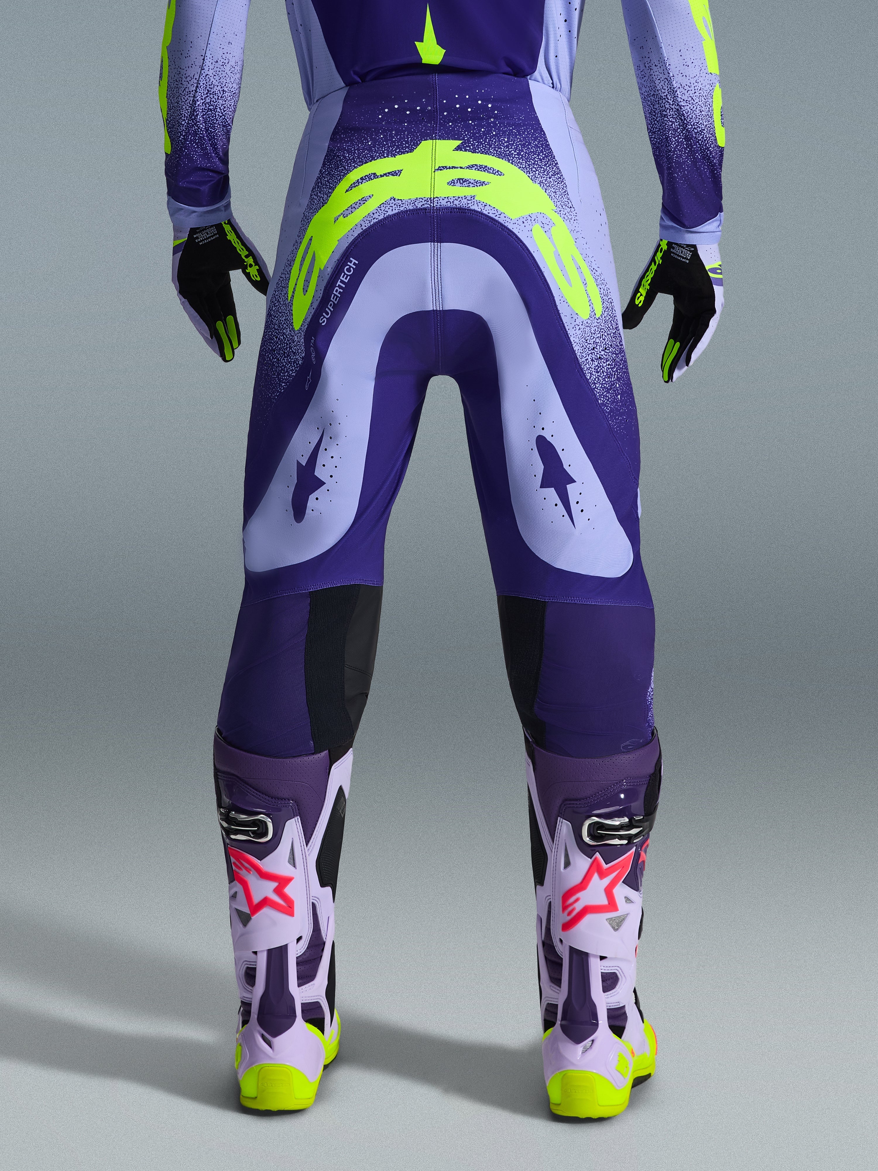 Alpinestars 2026 Supertech Scenz Hose, Motocross-Hose, Lila und Gelb Fluo, Rückansicht mit großem gelben Logo am Gesäß, getragen mit passendem Trikot, Handschuhen und Schutzstiefeln, athletische Passform für professionellen Motocross-Rennsport.