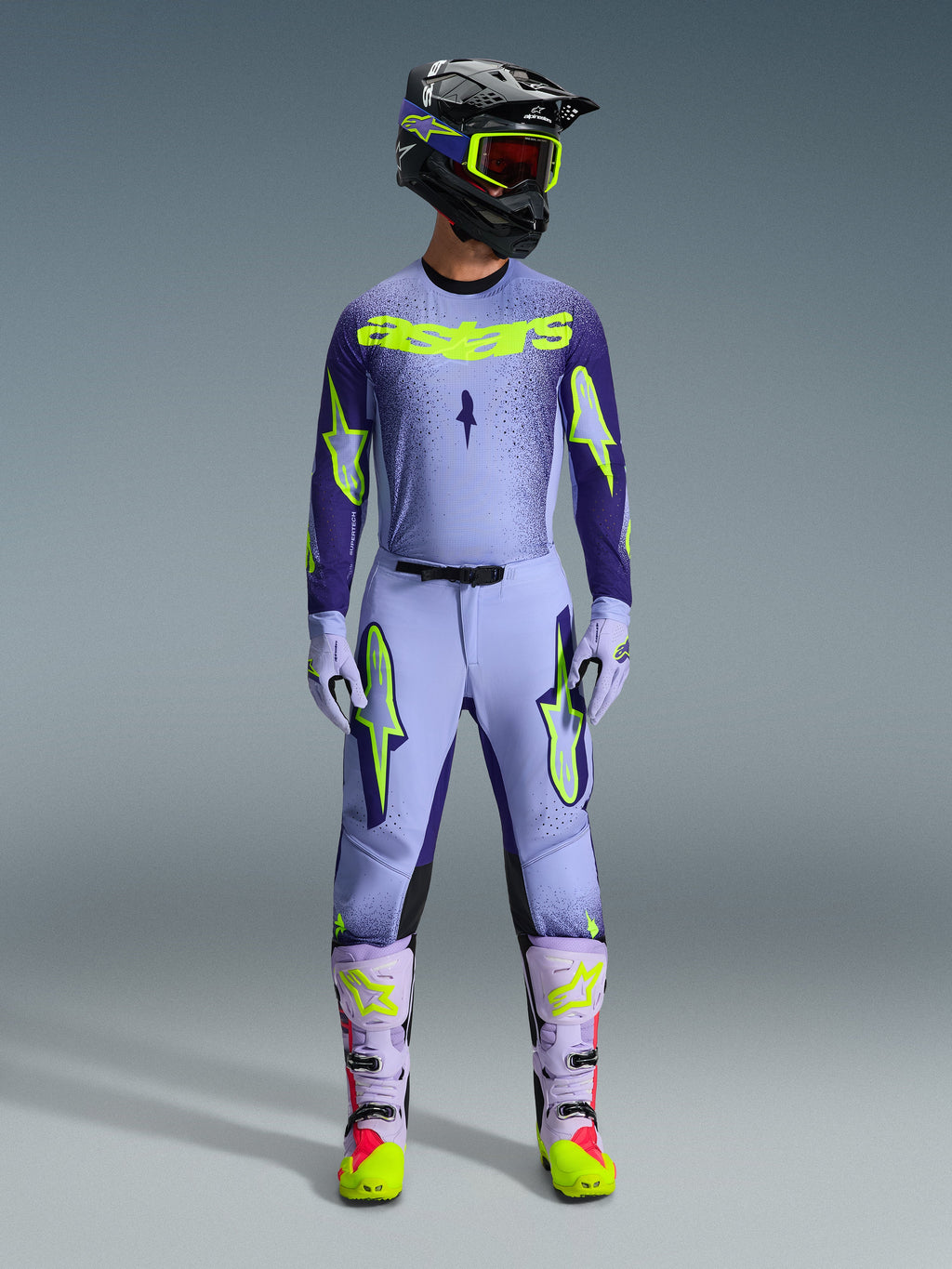 Alpinestars 2026 Supertech Scenz Gear Set, Motocross-Rennoutfit, Hellviolett und Neongelb-grün, komplette Schutzausrüstung, schwarzer Helm mit violetten Akzenten, Jersey mit auffälligem neongelbem Branding, passende Hose mit Stern-Grafiken, abgestimmte Stiefel mit rosa Highlights.