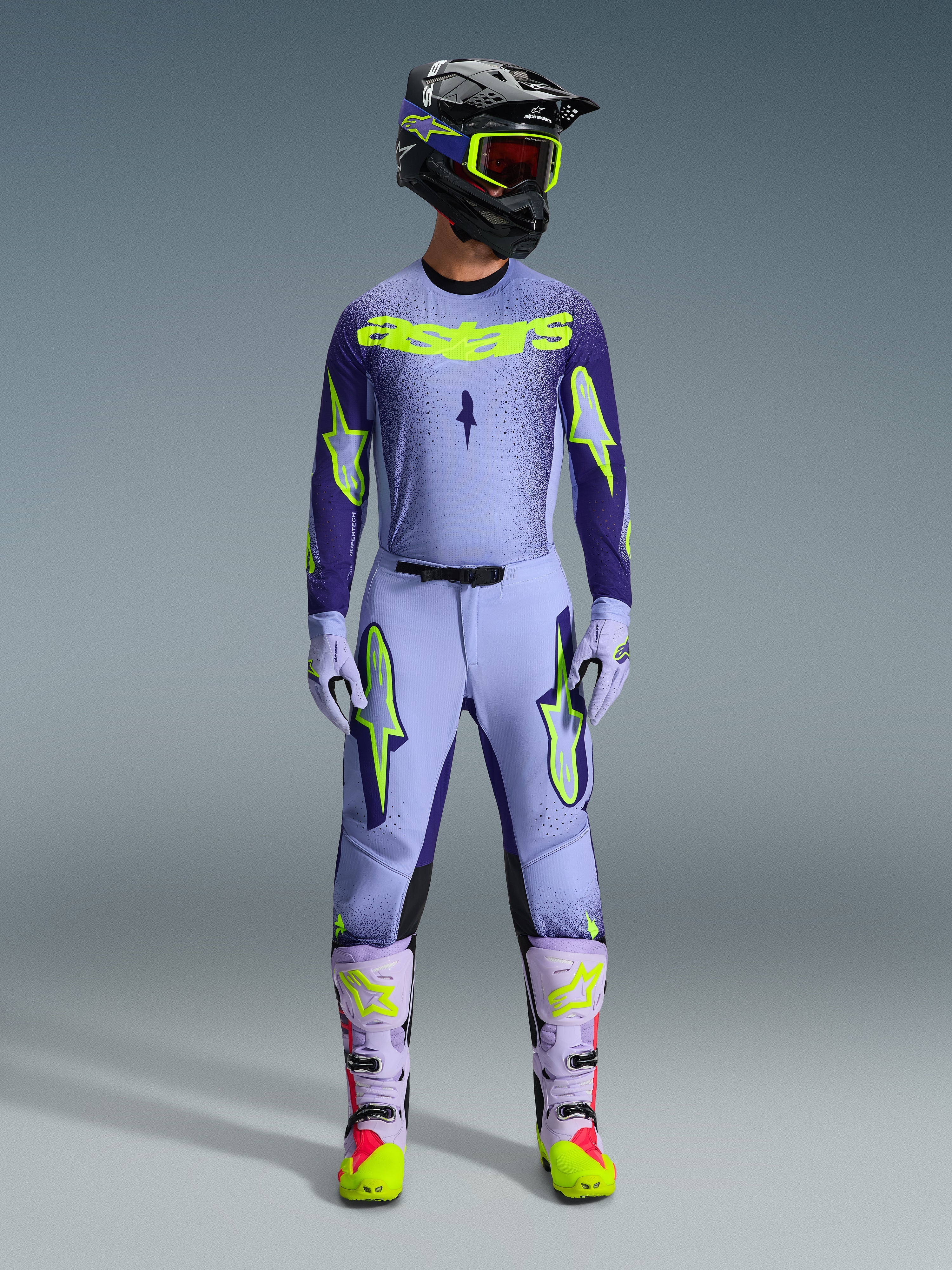 Alpinestars 2026 Supertech Scenz Gear Set, Motocross-Rennoutfit, Hellviolett und Neongelb-grün, komplette Schutzausrüstung, schwarzer Helm mit violetten Akzenten, Jersey mit auffälligem neongelbem Branding, passende Hose mit Stern-Grafiken, abgestimmte Stiefel mit rosa Highlights.
