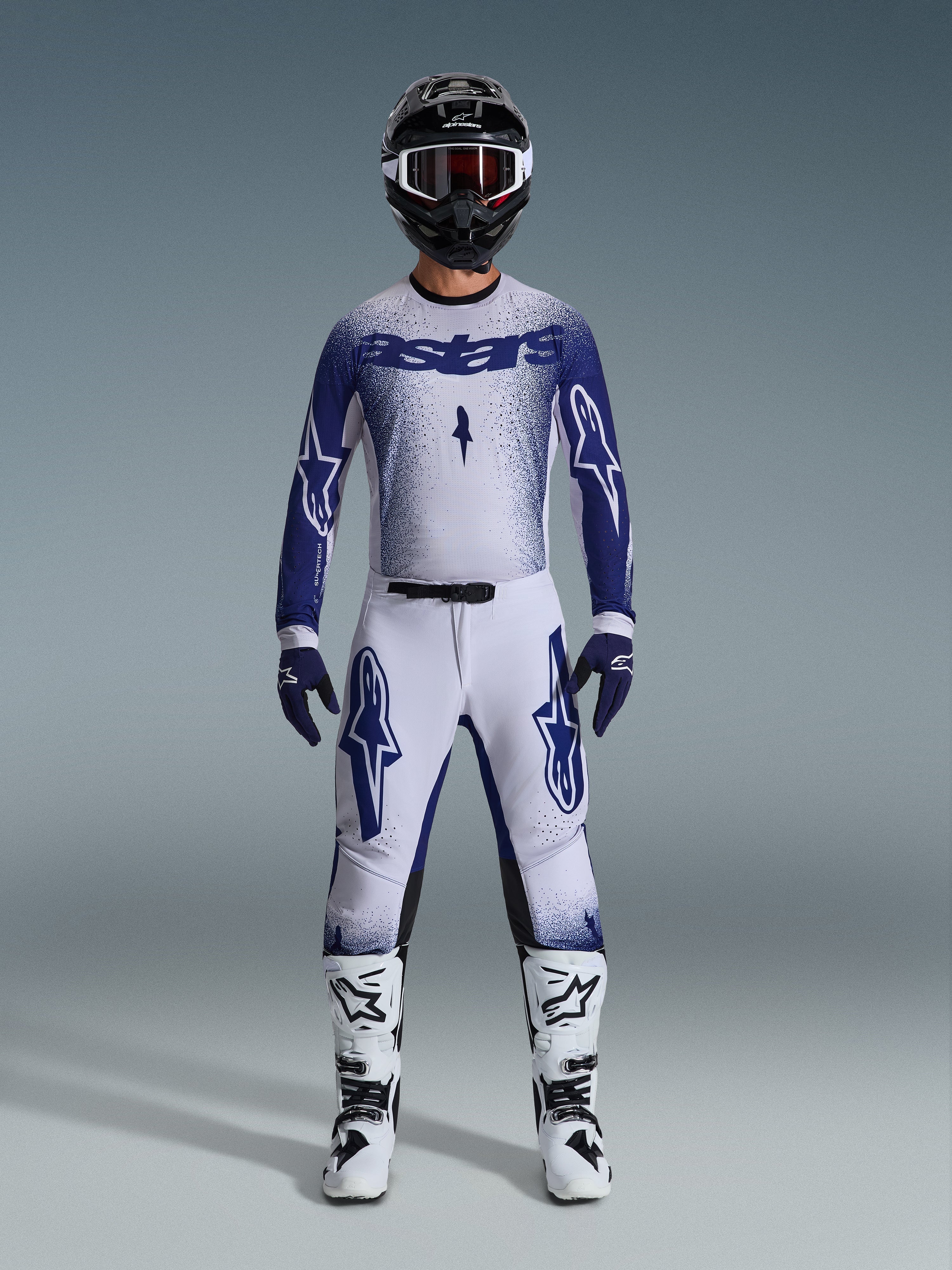 Motocross rider wearing Alpinestars 2026 Supertech Scenz gear set, Light Gray and Dark Blue, including a jersey and pants with matching blue gloves, white and black boots, and a black helmet with white goggles, coordinated outfit for professional off-road racing