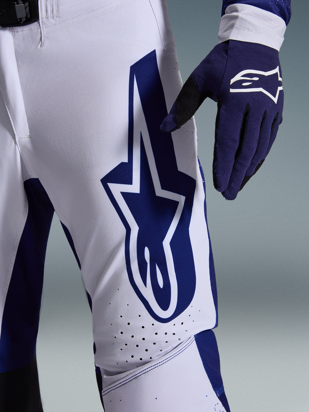 Alpinestars 2026 Supertech Scenz Pants, Motocross Pants, Light Gray and Dark Blue, close-up view of the thigh showing a large dark blue Alpinestars logo, perforated ventilation zones, and stretch rip-stop fabric, worn with a matching dark blue motocross glove