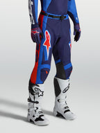 Alpinestars Racer Hunt broek, heren motorcrossbroek, blauw en vuurrood, duurzaam 600D polyweefsel met ergonomische voorgevormde racepasvorm, gedragen met witte motorlaarzen.