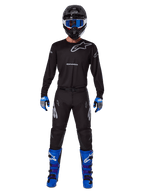 Pantalon Alpinestars 2025 Racer Graphite, pantalon de motocross, noir et gris, porté par un mannequin avec un maillot noir assorti, des bottes bleues et violettes et un masque bleu, tenue de course tout-terrain complète avec logo Alpinestars