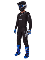 Pilote de motocross portant un équipement complet Alpinestars, pantalon Racer Graphite 2025 noir et gris, maillot noir assorti avec logos gris, casque noir avec lunettes bleues, bottes bleu vif et violet, et gants bleus et gris, tenue coordonnée pour la course tout-terrain
