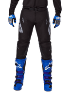 Pantalon Alpinestars Racer Graphite 2025, pantalon de motocross, noir et gris, porté avec des gants bleus et gris et des bottes bleues, avec marquage Astars argenté sur les cuisses et genoux renforcés, design sportif pour la performance tout-terrain