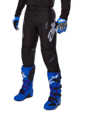 Pantalon Alpinestars Racer Graphite 2025, pantalon de motocross noir avec graphismes blancs et gris, porté avec des bottes de motocross bleu vif et des gants bleus assortis, tissu technique durable pour la conduite tout-terrain