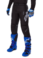 Pantalon Alpinestars Racer Graphite 2025, pantalon de motocross noir avec graphismes blancs et gris, porté avec des bottes de motocross bleu vif et des gants bleus assortis, tissu technique durable pour la conduite tout-terrain
