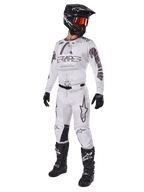 Alpinestars 2025 Racer Hollow broek, motorcross broek, lichtbruine camouflage, getoond als onderdeel van een complete race-uitrusting met een wit shirt, zwarte helm en zwarte laarzen, voorzien van versterkte zwarte kniepanelen en een subtiel camouflage-ontwerp.