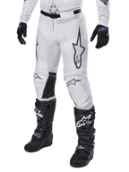 Alpinestars 2025 Racer Hollow Pants, motorcross broek, wit en grijs camo, met grote zwart omijnde sterlogo's en zwarte leren binnenkniepanelen, gedragen met zwarte motorcrosslaarzen en handschoenen, duurzaam ontwerp voor terreinrijden