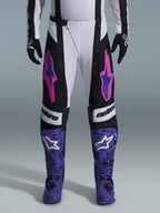 Alpinestars 2026 Techstar Knif Hose, Motocross-Hose, Schwarz, Weiß und Lila mit pinkfarbenen und blauen Akzenten, getragen mit lila Rennstiefeln und schwarzen Handschuhen, verfügt über einen ergonomischen Rennschnitt und markante Logo-Grafiken für Offroad-Performance