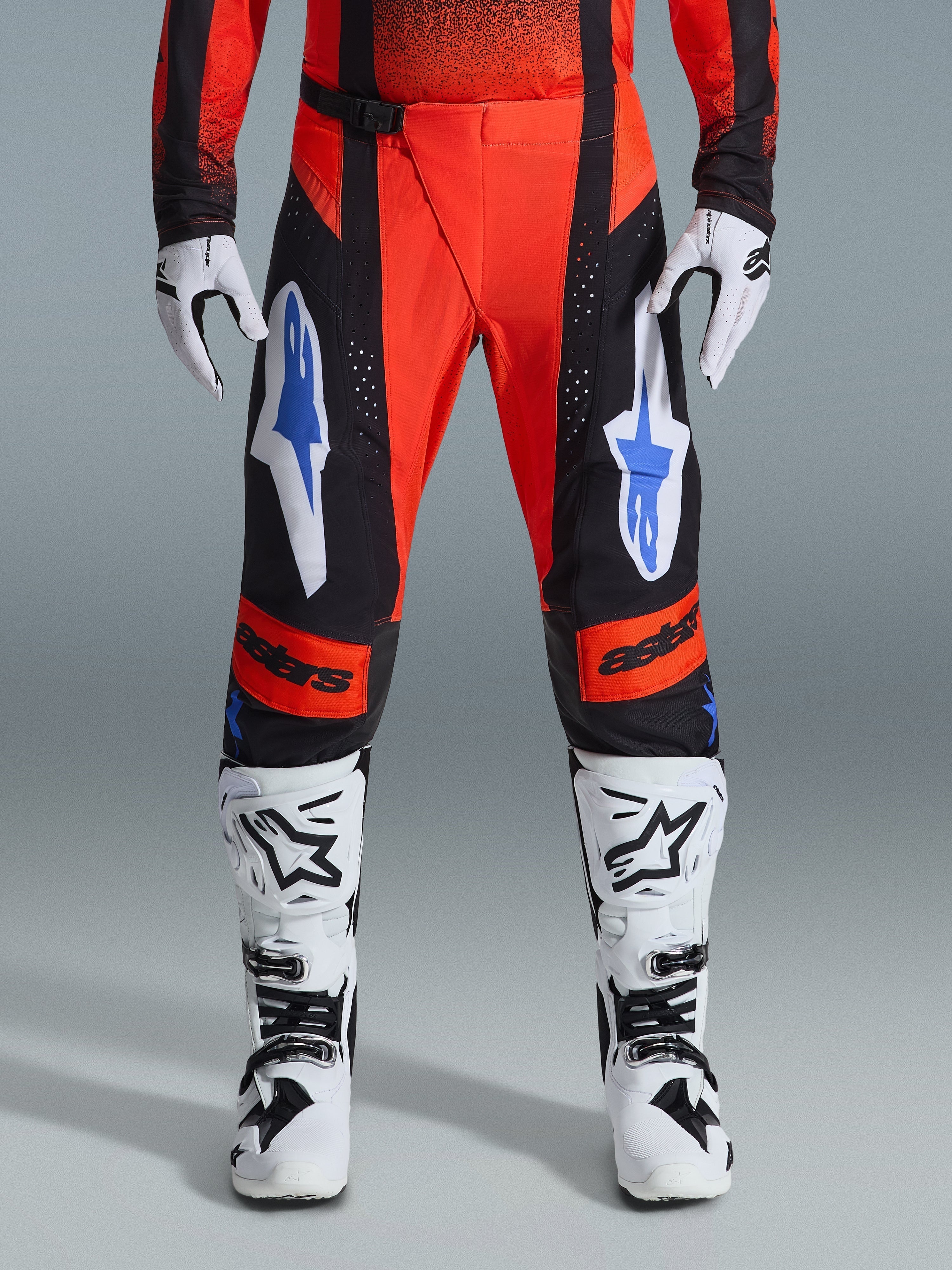 Alpinestars 2026 Techstar Knif crossbroek, motorcross broek, oranje, zwart en UCLA blauw, gedragen met bijpassende jersey, witte en zwarte motorcrosslaarzen en handschoenen, met opvallende branding op de dijen en knieën, sportieve racepasvorm voor off-road rijden