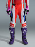 Alpinestars 2026 Techstar Knif Broek, motorcrossbroek in oranje fluo en lichtgrijs met zwarte accenten, getoond met paarse motorcrosslaarzen en handschoenen, voorzien van blauwe ster-logo's en multi-paneel constructie voor off-road prestaties
