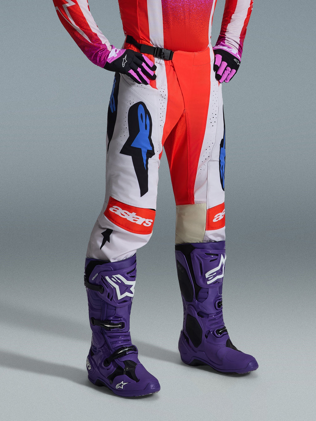 Alpinestars 2026 Techstar Knif Broek, motorcrossbroek, wit en helderrood met blauwe grafische accenten, met een "astars"-logo op de knie, gedragen met paarse laarzen en zwart-roze handschoenen, professionele off-road race-uitrusting.