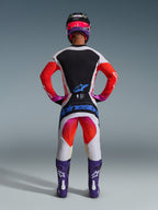 Motorcrosser van achteren in 2026 Techstar Knif Broek en bijpassende jersey, lichtgrijs en zwart ontwerp met feloranje en paarse accenten, blauwe Alpinestars-branding op het zitvlak, paarse laarzen en helm, professionele off-road racekledingset