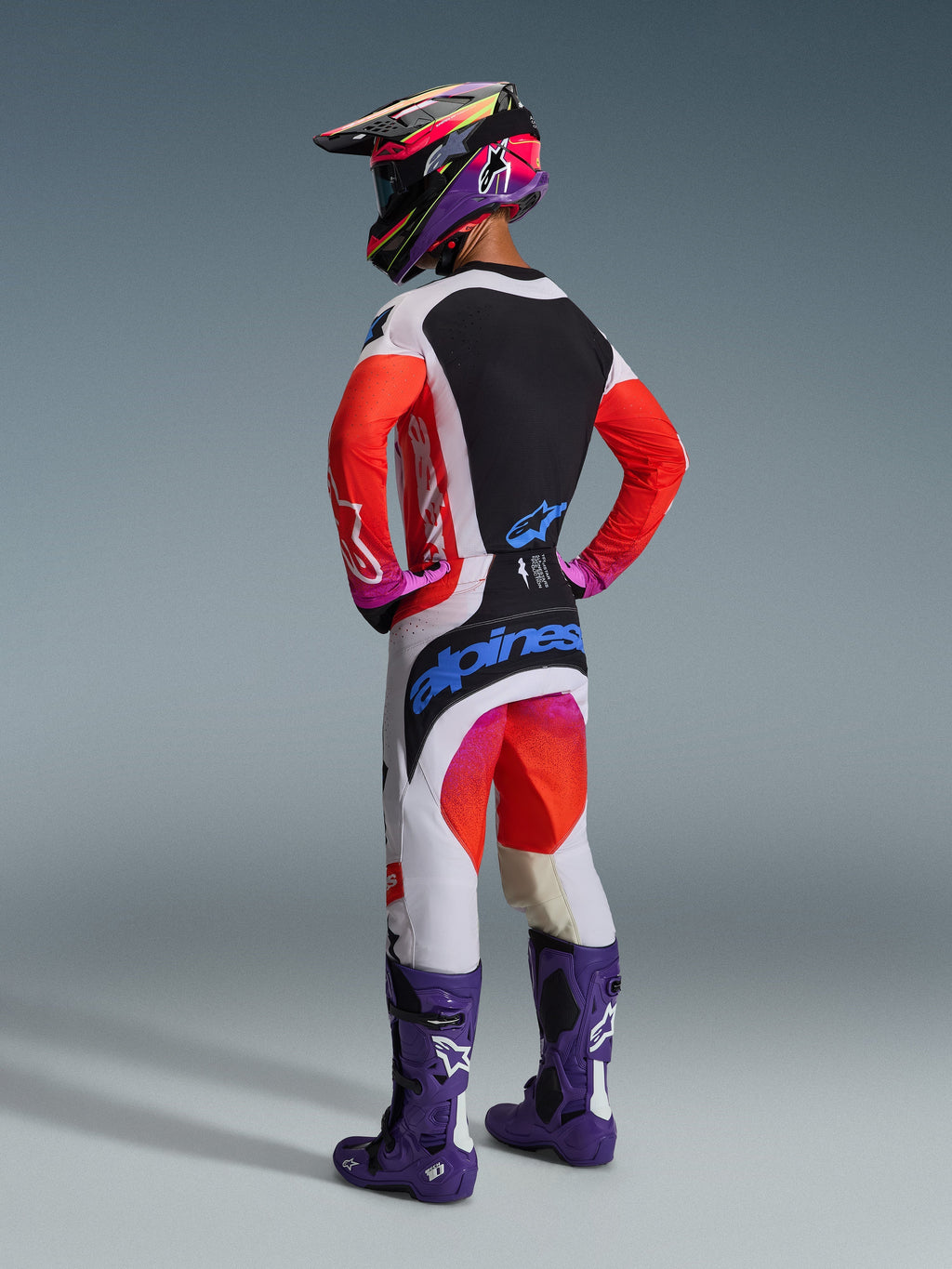 Motorcrosser van achteren gezien in volledige race-uitrusting, Alpinestars 2026 Techstar Knif Broek in lichtgrijs en fluo oranje met blauwe merkaccenten, bijpassende jersey met oranje mouwen, paarse Tech 10 laarzen en een kleurrijke racehelm, gecoördineerde professionele off-road outfit