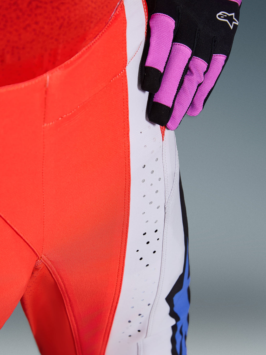 Alpinestars 2026 Techstar Knif Pants, motorcrossbroek, oranje fluo en lichtgrijs met blauwe accenten, close-up van lasergesneden ventilatieperforaties op het dijbeen, gedragen met een zwart-paarse motorcrosshandschoen