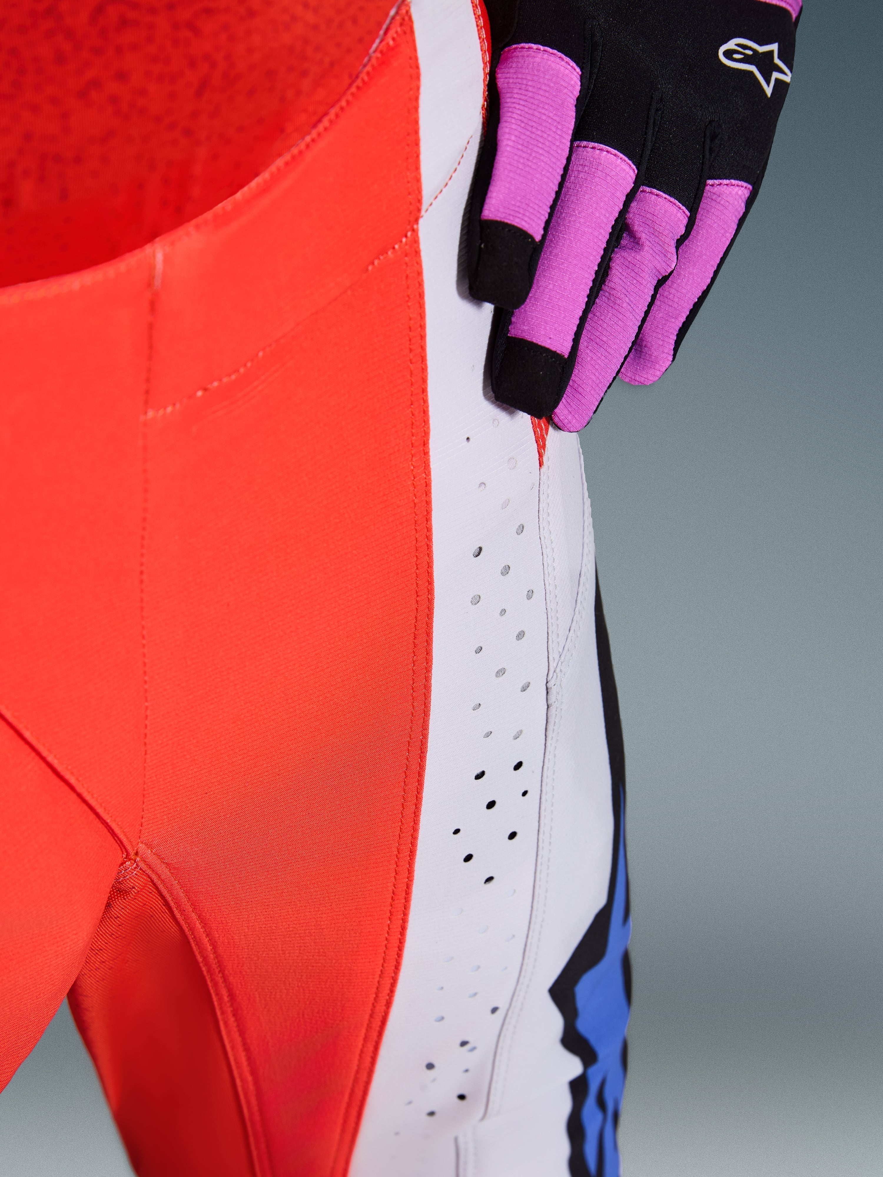 Alpinestars 2026 Techstar Knif Pants, motorcrossbroek, oranje fluo en lichtgrijs met blauwe accenten, close-up van lasergesneden ventilatieperforaties op het dijbeen, gedragen met een zwart-paarse motorcrosshandschoen
