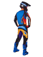 Motocross-Fahrer von hinten in der Alpinestars 2025 Fluid Wurx Hose, mehrfarbiges Design mit blauen, orangefarbenen, gelben und lila Akzenten, kombiniert mit einem passenden Trikot, leuchtend blauen Stiefeln und schwarzem Helm, komplette Offroad-Rennausrüstung.