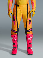 Alpinestars 2026 Techstar Nomur Hose, Motocross-Hose, Orange, Gelb und Pink, getragen mit passenden gelben Handschuhen und leuchtend pinkfarbenen Rennstiefeln, mit schwarzen Einsätzen und auffälligem pinkfarbenem Alpinestars-Branding, technische Offroad-Rennausrüstung