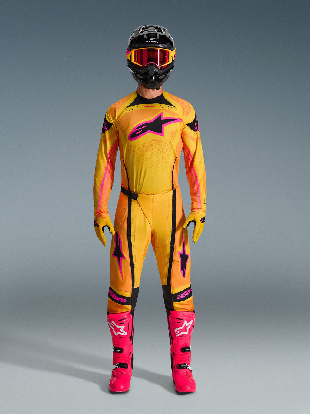 Motocross-Fahrer in vollständiger Alpinestars 2026 Techstar Nomur Ausrüstung, Trikot und Hose in Orange, Gelb und Pink, passenden gelben und pinken Handschuhen, leuchtend pinken Rennstiefeln und schwarzem Helm mit pinker Brille, koordiniertes Hochleistungs-Motocross-Outfit