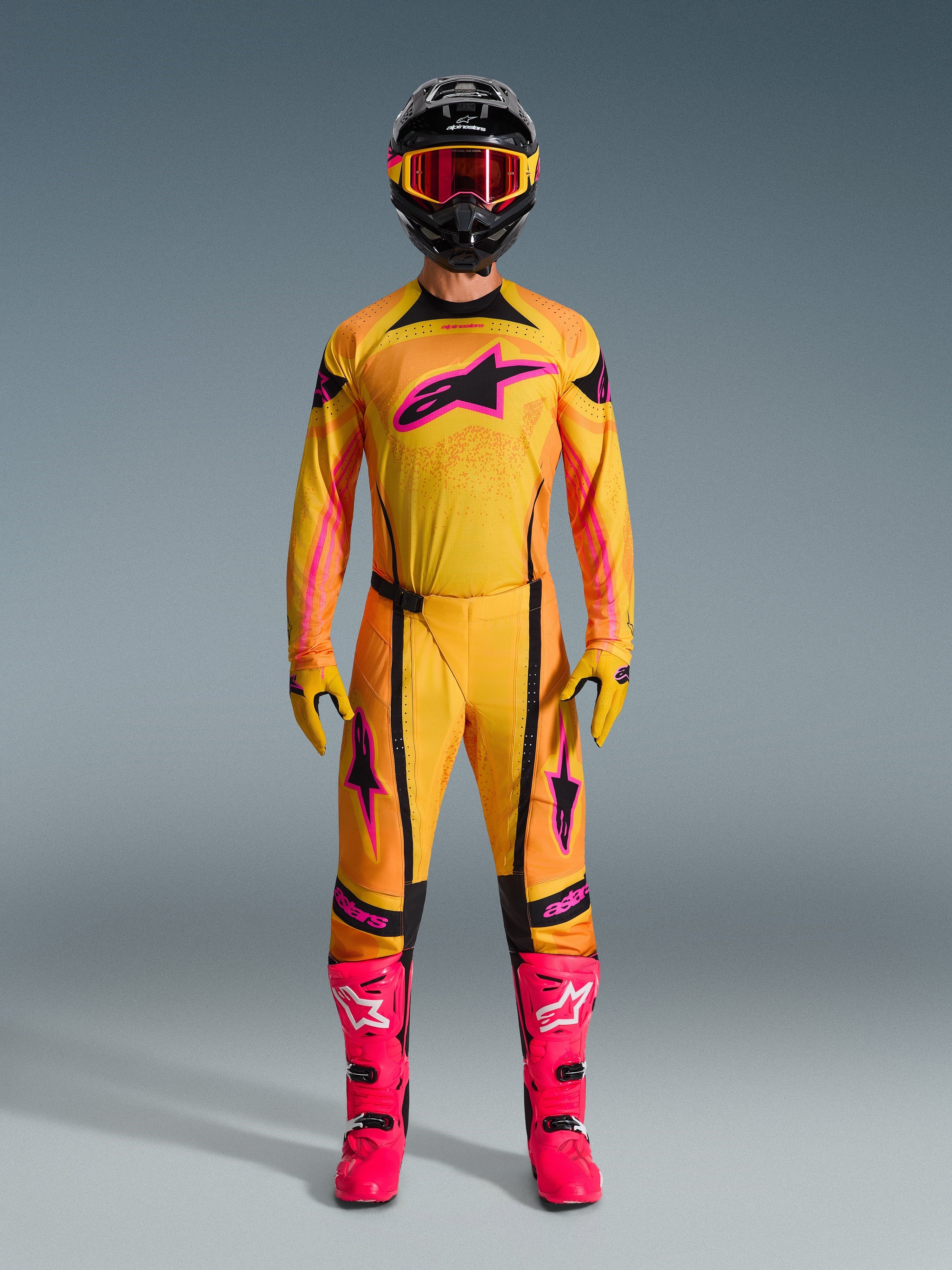 Motocross-Fahrer in vollständiger Alpinestars 2026 Techstar Nomur Ausrüstung, Trikot und Hose in Orange, Gelb und Pink, passenden gelben und pinken Handschuhen, leuchtend pinken Rennstiefeln und schwarzem Helm mit pinker Brille, koordiniertes Hochleistungs-Motocross-Outfit