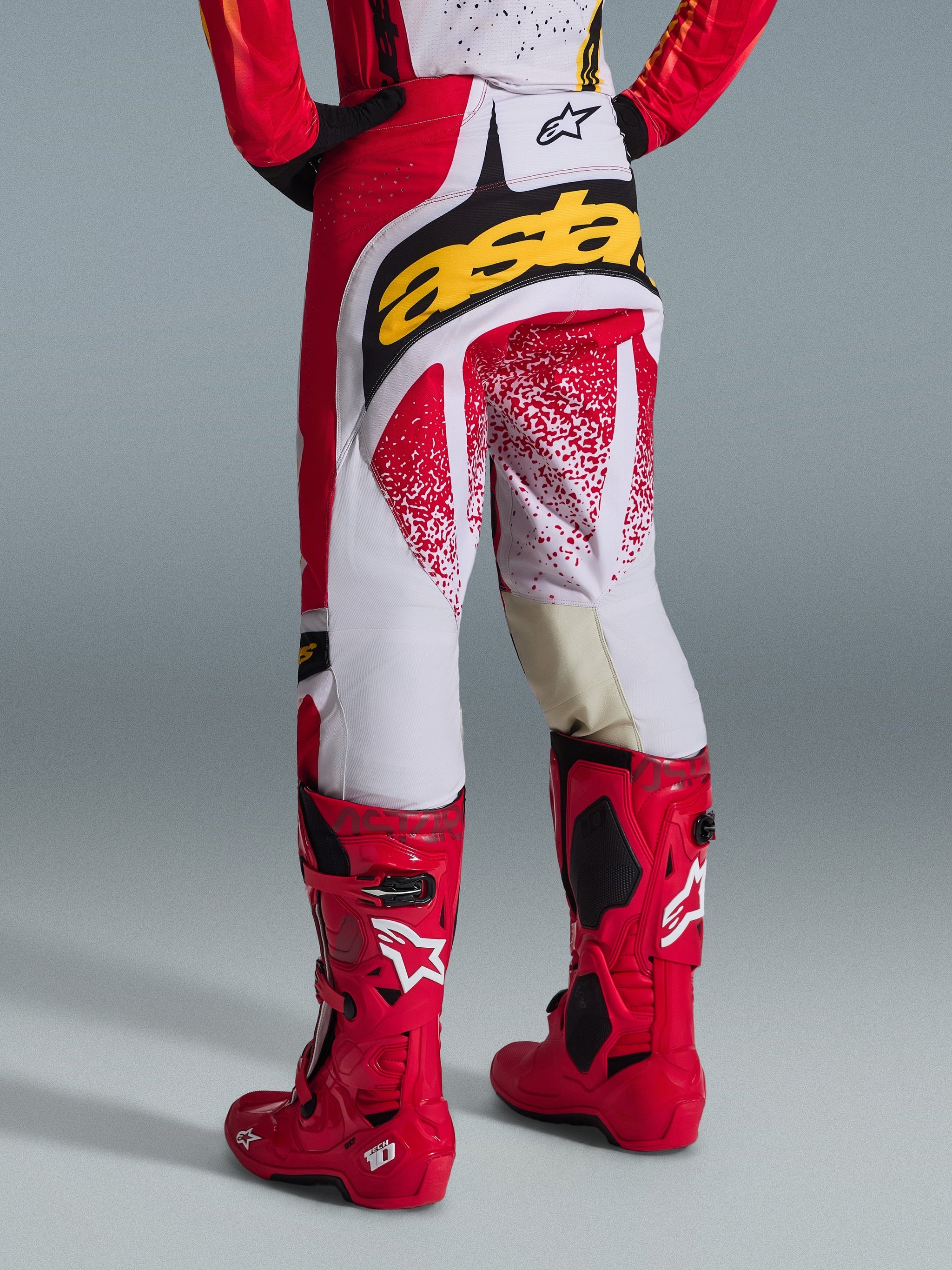 Vue arrière d'un pilote de motocross portant un pantalon Alpinestars Techstar Nomur 2026 rouge et blanc avec un logo jaune vif sur l'assise, associé à des bottes de motocross Tech 10 rouge vif, photo de studio sur fond gris.