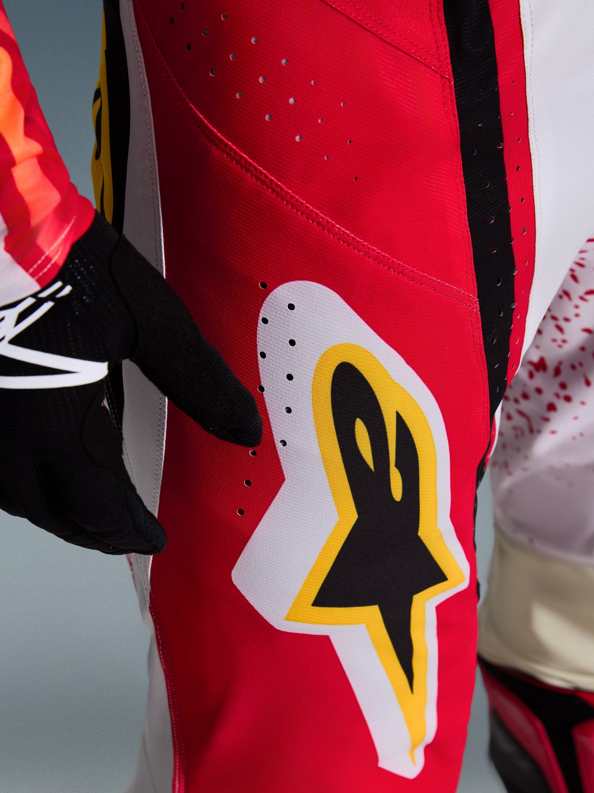 Alpinestars 2026 Techstar Nomur Pants, pantalon de motocross, rouge avec accents blancs et jaunes, gros plan sur la zone de la cuisse présentant des perforations de ventilation laser et un grand logo en étoile, associé à des gants noirs et un maillot coloré pour la performance tout-terrain