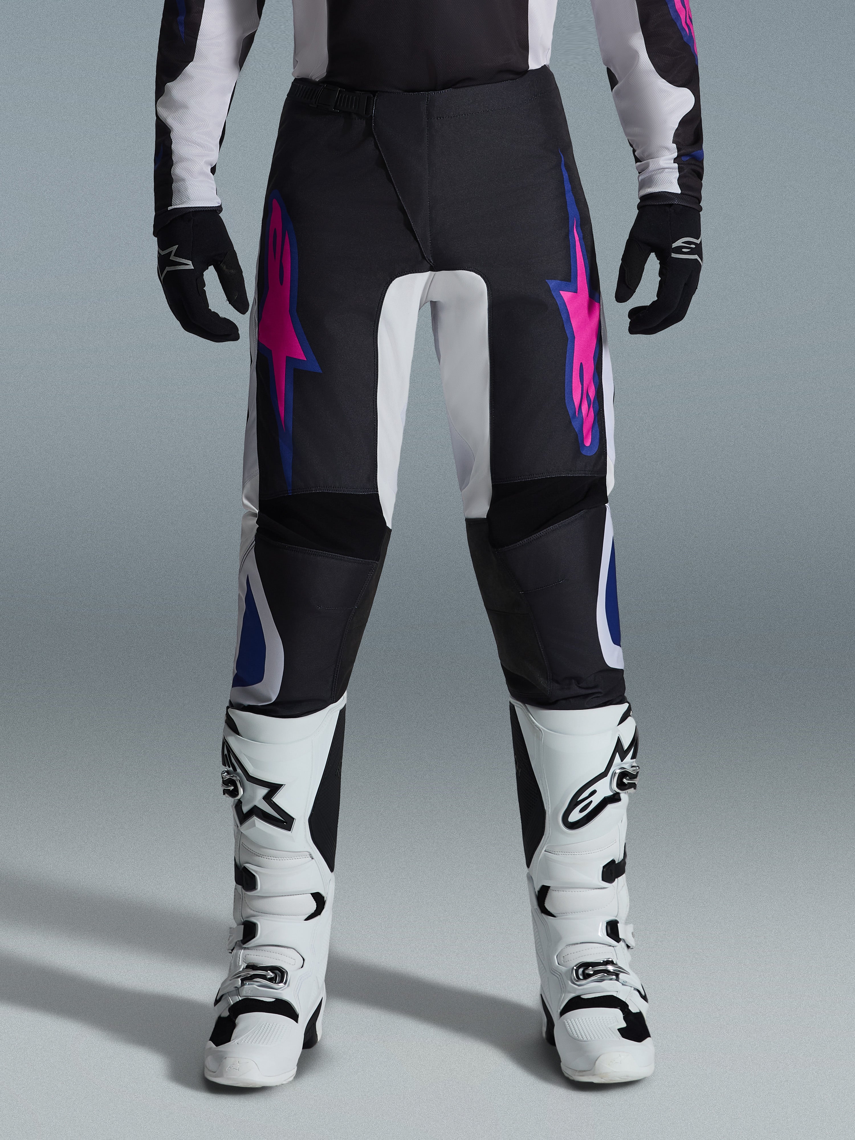Alpinestars 2026 Fluid Grid Pants, pantalon de motocross, noir avec motifs d'étoiles violets et bleus, porté avec des bottes de motocross blanches et des gants noirs, avec empiècements renforcés et coupe sportive tout-terrain, fond gris