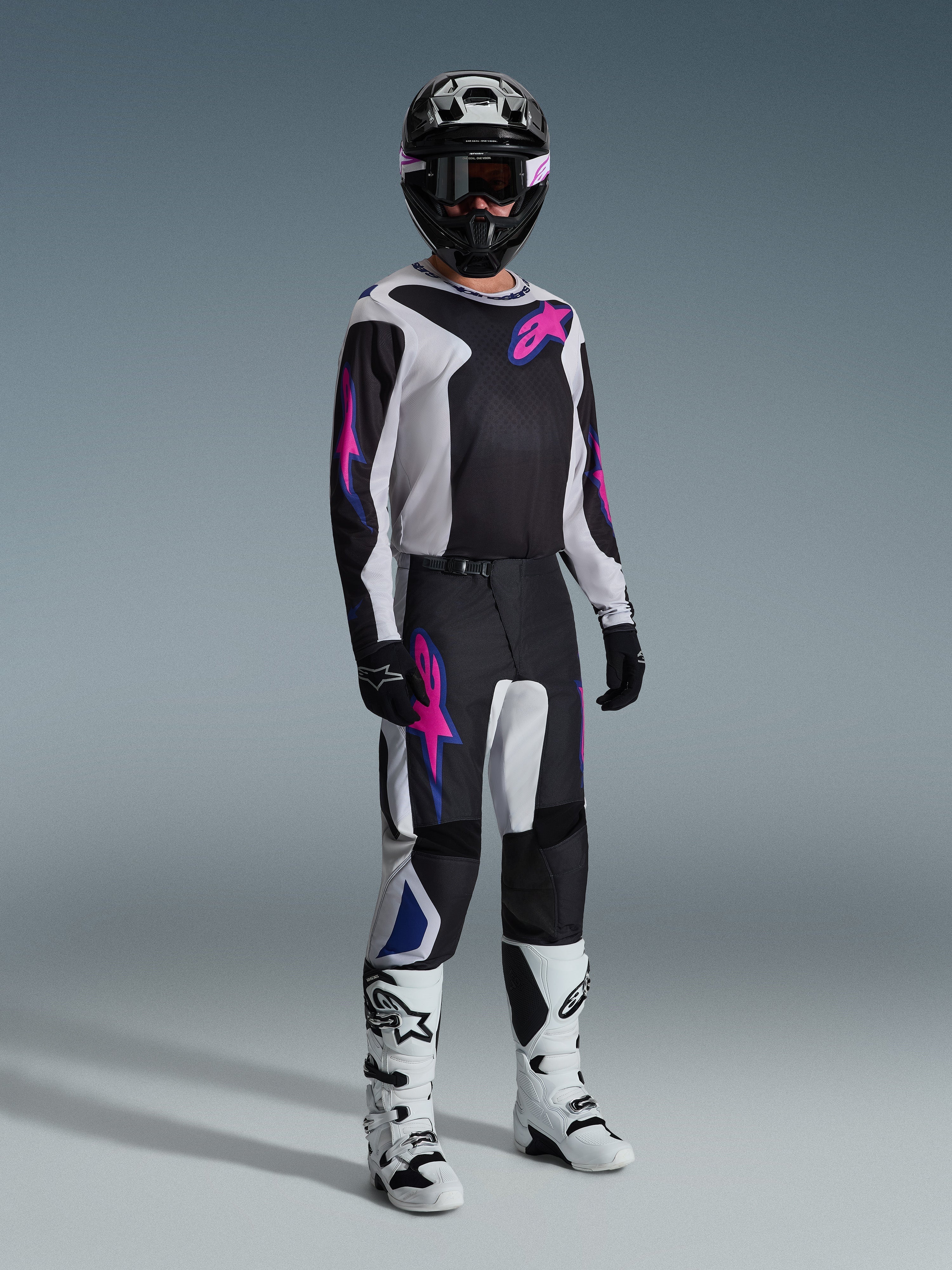 Motocross rider wearing complete racing gear, Alpinestars 2026 Fluid Grid Pants in Black Light Gray and Purple, matching jersey with pink and blue graphics, black helmet and goggles, white and black boots, and black gloves, coordinated outfit for off-road racing