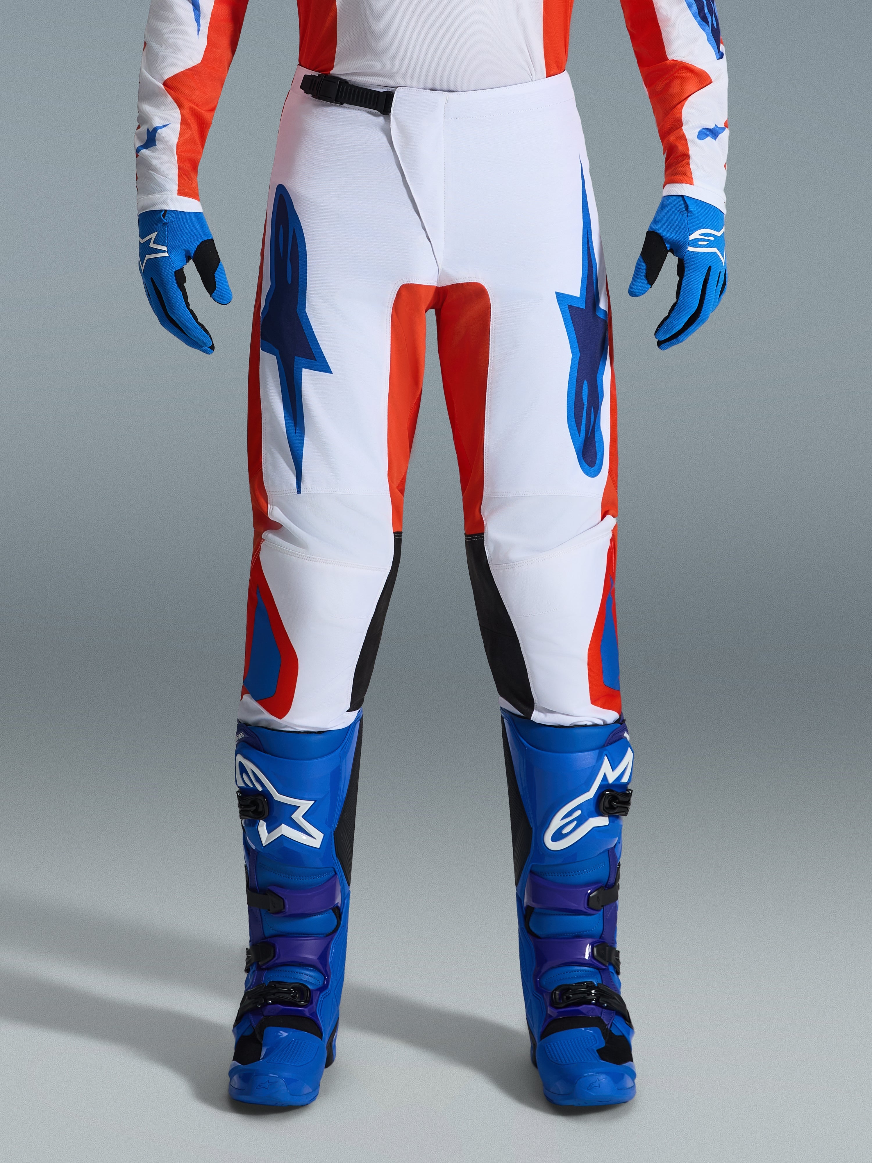 Alpinestars 2026 Fluid Grid Pants, pantalon de motocross, orange et bleu UCLA, porté avec des bottes et des gants bleus assortis, avec un châssis blanc, des logos bleus audacieux et des accents orange, design ergonomique pour la performance tout-terrain