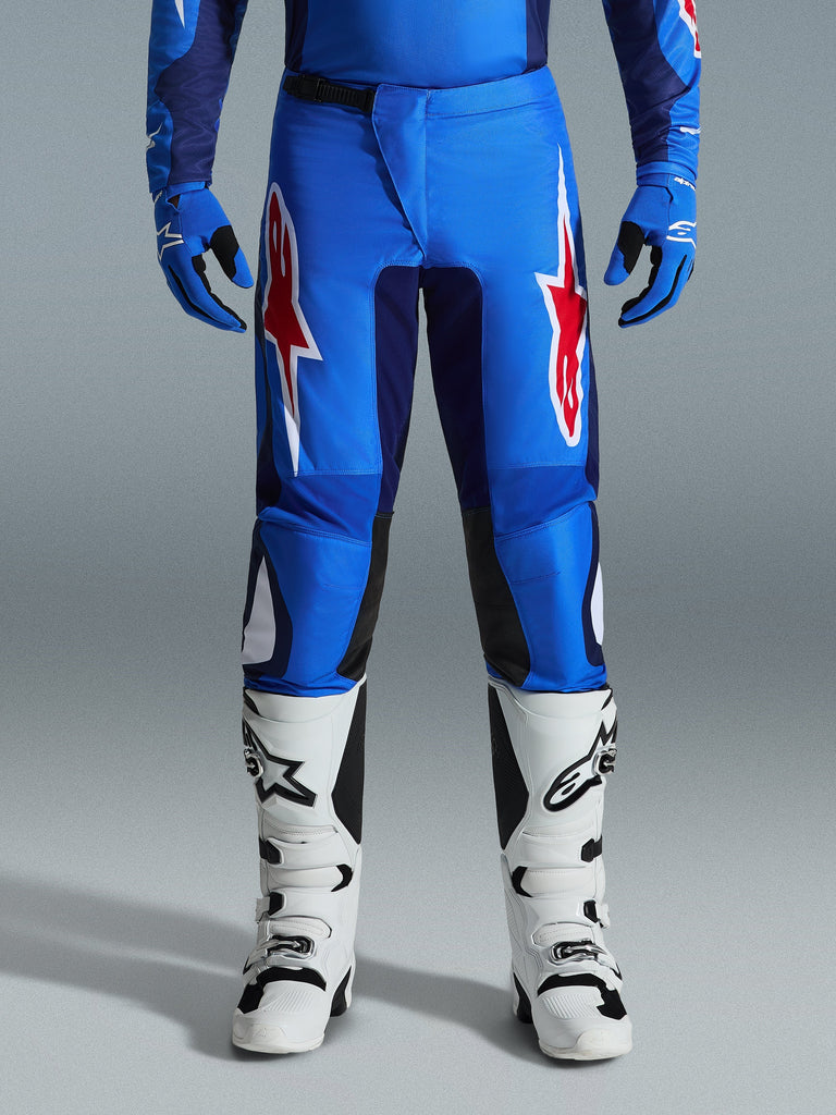 Alpinestars 2026 Fluid Grid Pants, motorcrossbroek, blauw en rood, met opvallende rode en witte sterlogo's op de dijen, gedragen met bijpassende blauwe handschoenen en witte off-road laarzen, sportieve pasvorm met duurzame panelen voor racen.