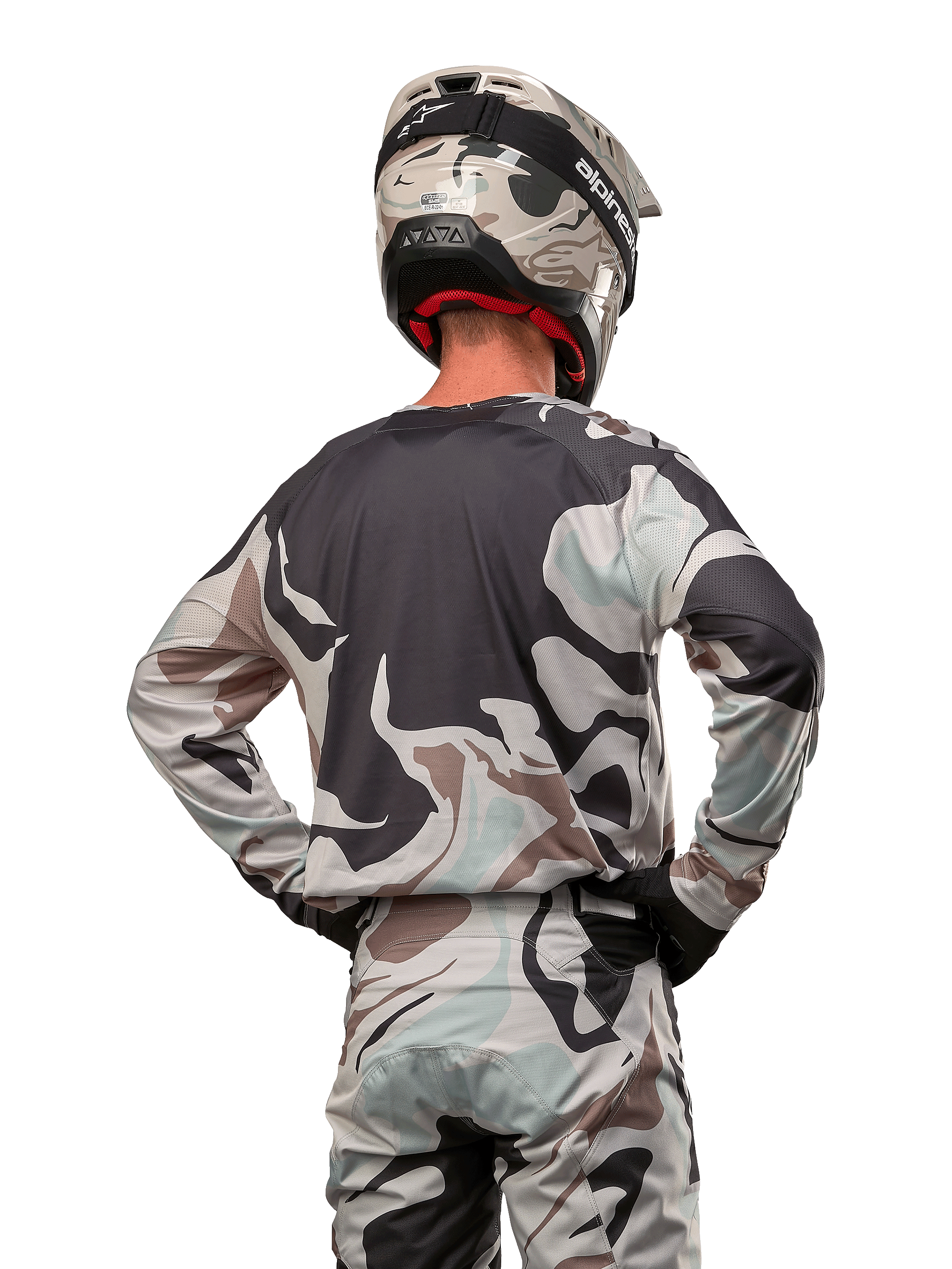 Pilote de motocross vu de dos portant le maillot Alpinestars Racer Tactical 2024 en Iron Camo Dust Gray, pantalon et casque camouflage assortis, mains sur les hanches, équipement de course tout-terrain coordonné.