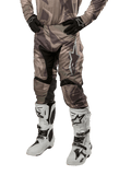 Alpinestars 2024 Racer Tactical Pants, pantalon de motocross, camouflage vert militaire et marron, porté avec un maillot camouflage assorti, des gants noirs et des bottes blanches avec des logos étoiles, doté de panneaux extensibles noirs et d'un design ergonomique, tenue tout-terrain sur fond blanc