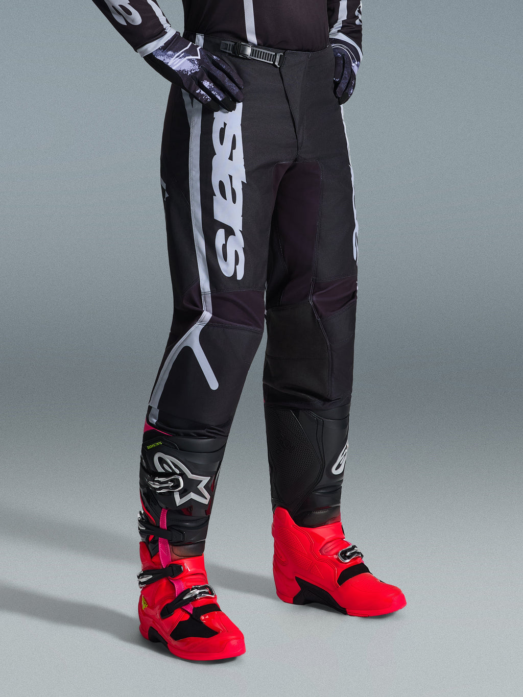 Alpinestars 2026 Fluid Apex Pants, Motocross Pants, Black and Gray, worn with vibrant pink and red boots and matching black and white jersey, featuring bold vertical Alpinestars branding on the thigh, reinforced panels, and ratchet waist closure for off-road performance.