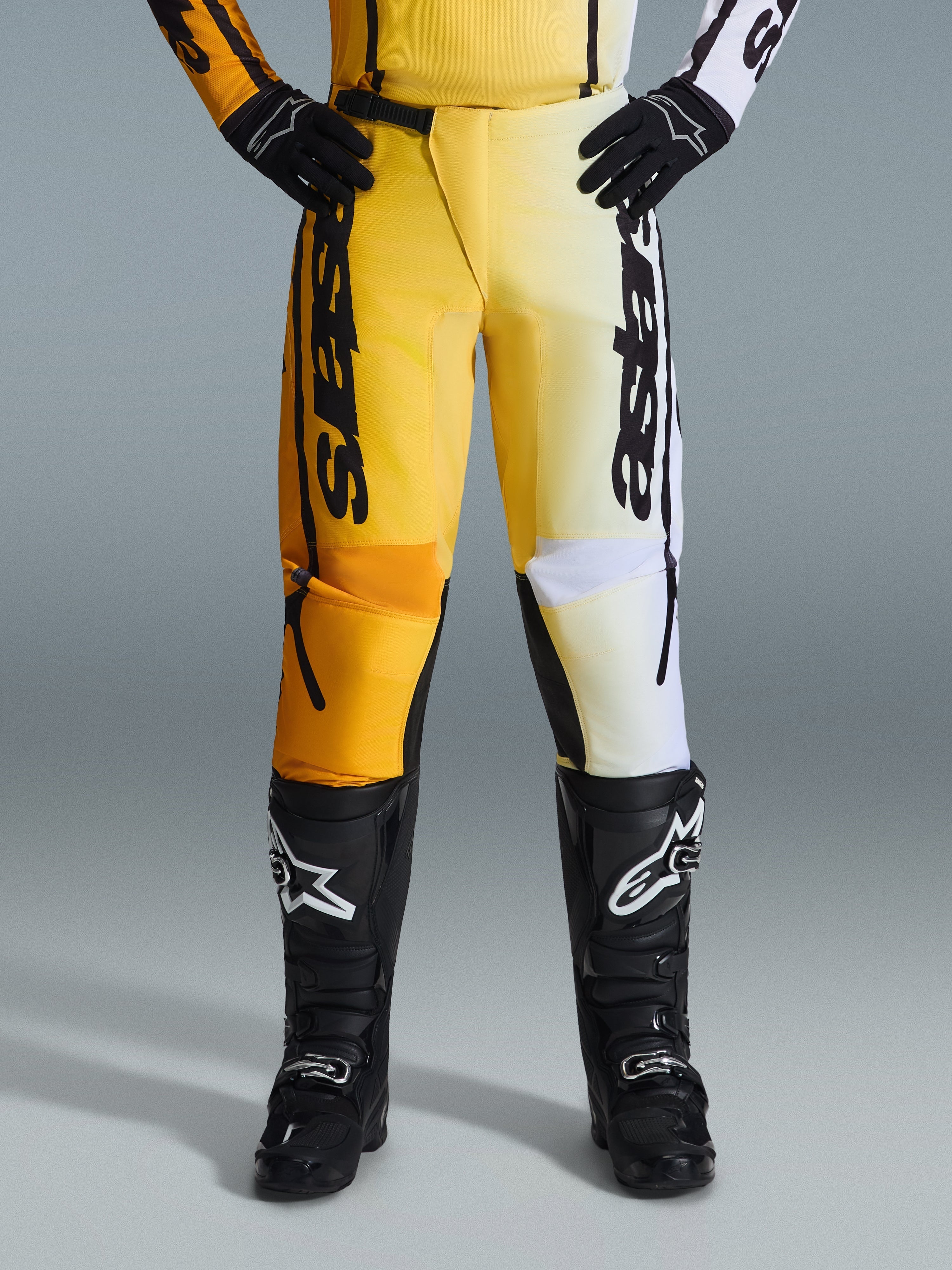 Alpinestars 2026 Fluid Apex broek, motorcrossbroek, geel en zwart, tweekleurig ontwerp met gele en witte panelen, opvallende verticale 'astars' branding, gedragen met zwarte motorcrosslaarzen en handschoenen, performance kleding voor off-road rijden
