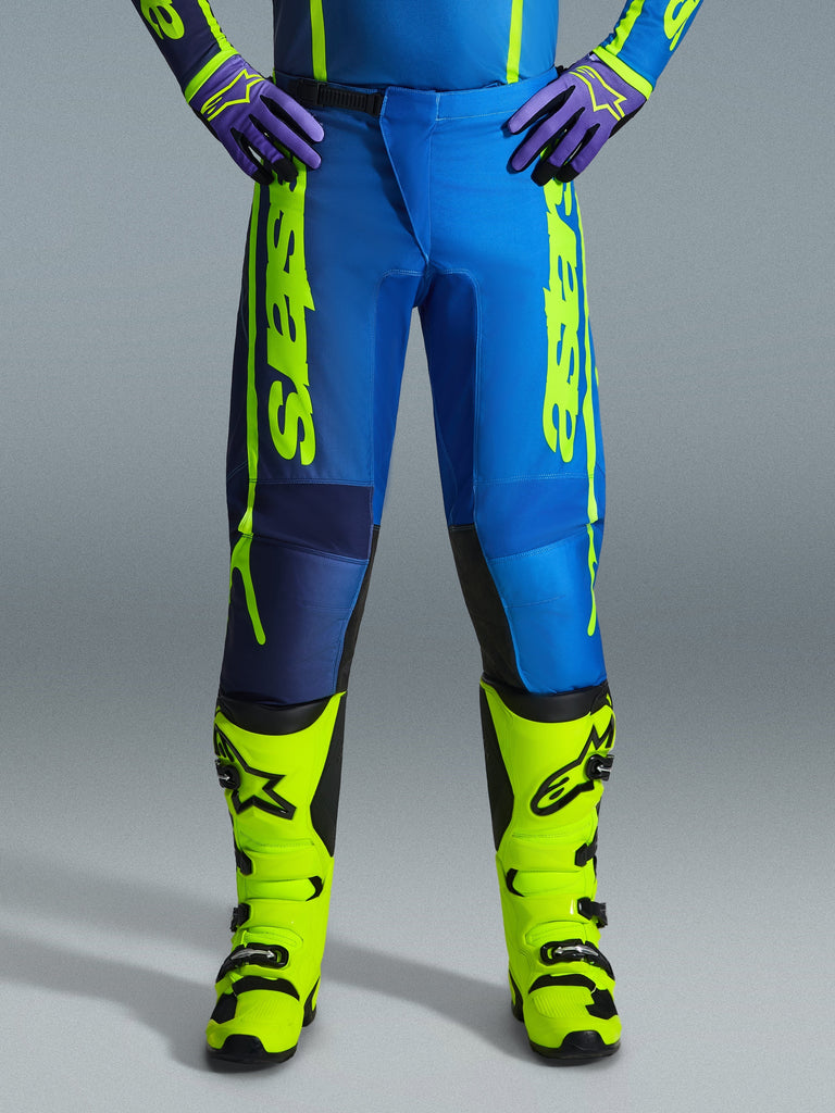 Alpinestars 2026 Fluid Apex crossbroek, motorcross broek, blauw en fluo geel, gedragen met bijpassende fluo gele laarzen en paarse handschoenen, opvallende gele branding aan de zijkanten, ergonomisch ontwerp voor off-road racing