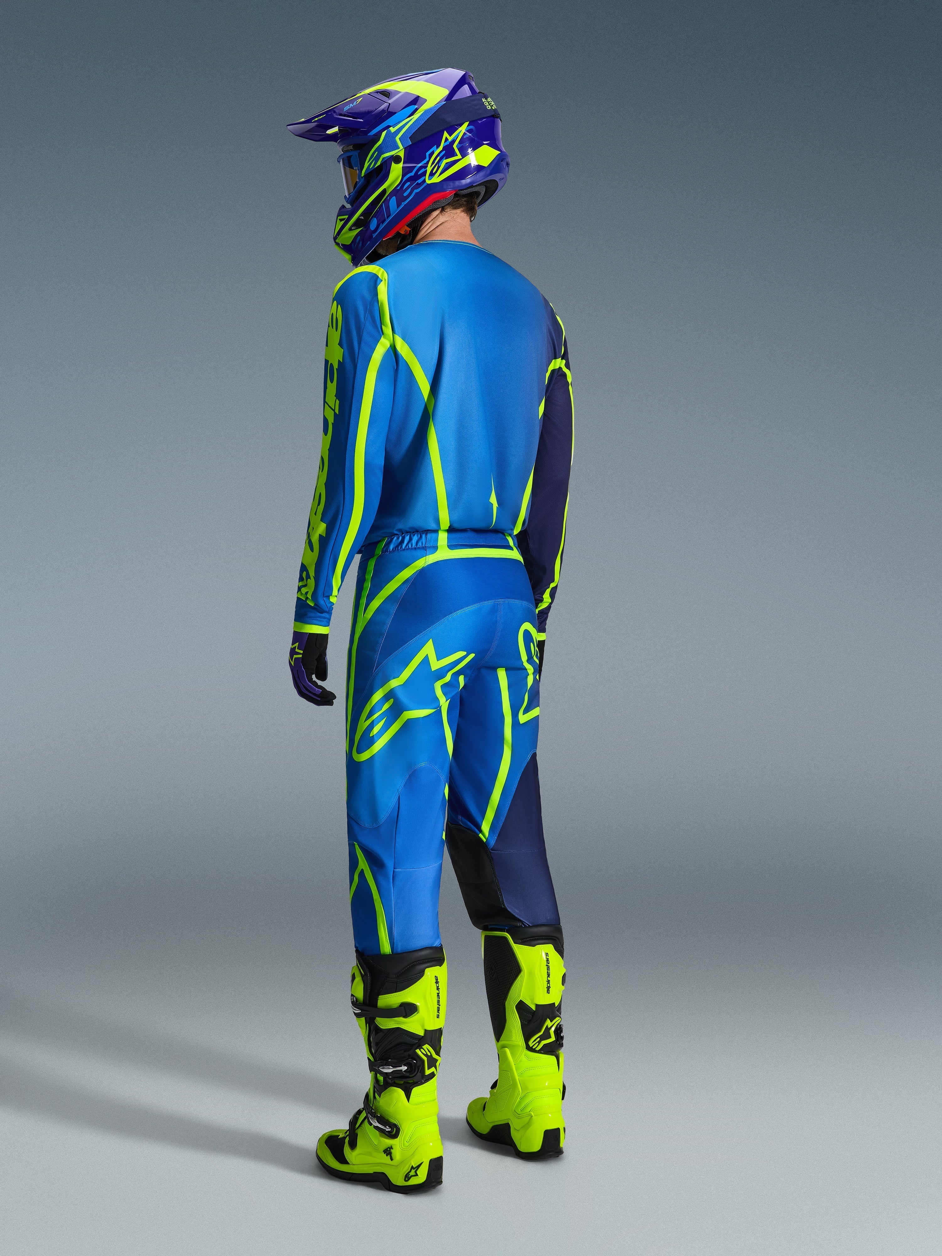 Motocross rider from a rear view wearing 2026 Alpinestars Fluid Apex Pants in Blue and Yellow Fluo, matching blue and yellow jersey and helmet, and bright neon yellow boots, coordinated racing gear for off-road performance.