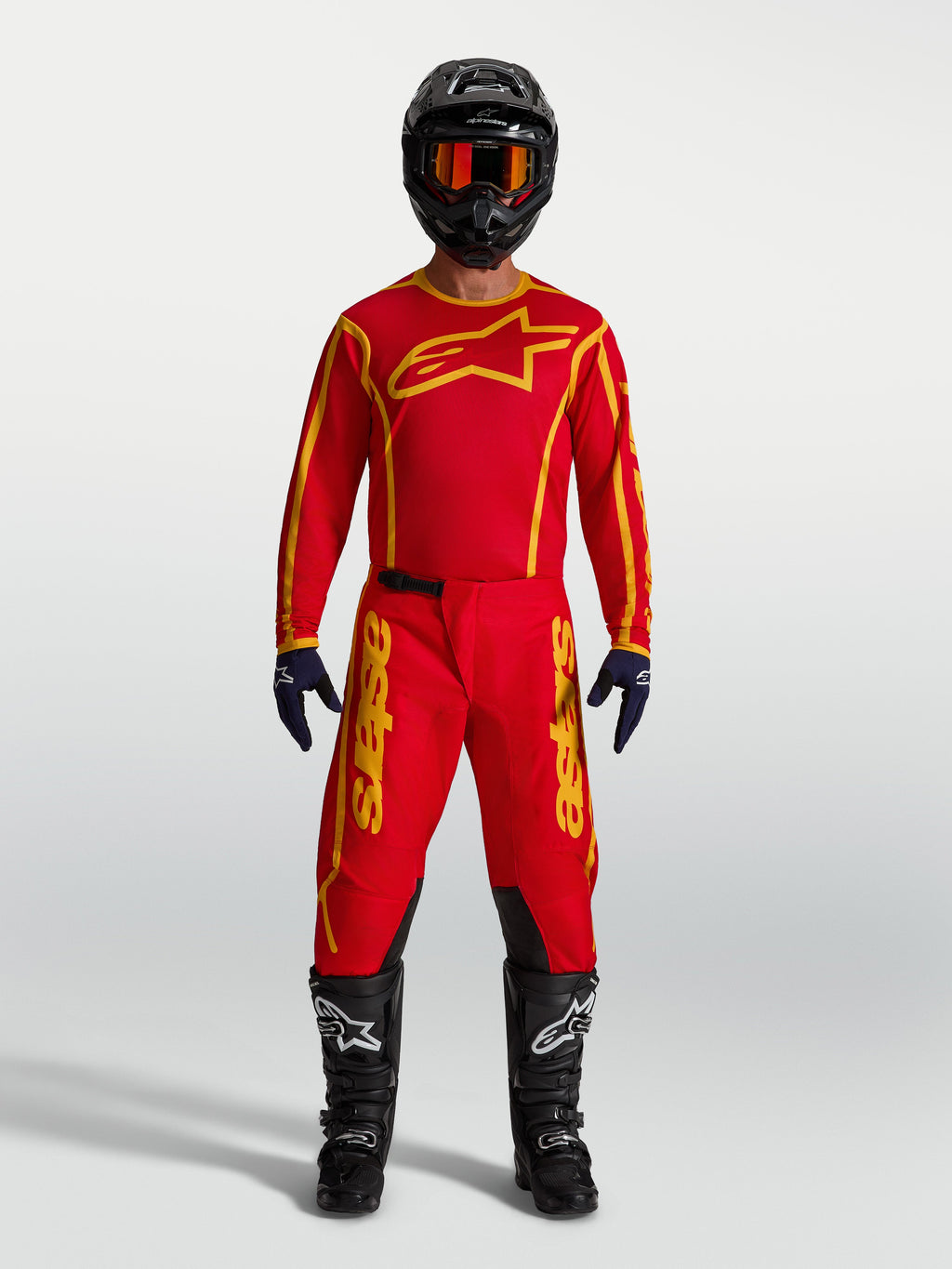 Alpinestars Fluid Apex V2 broek, heren motocrossuitrusting in rood en goud met jersey, zwarte helm en motorlaarzen.