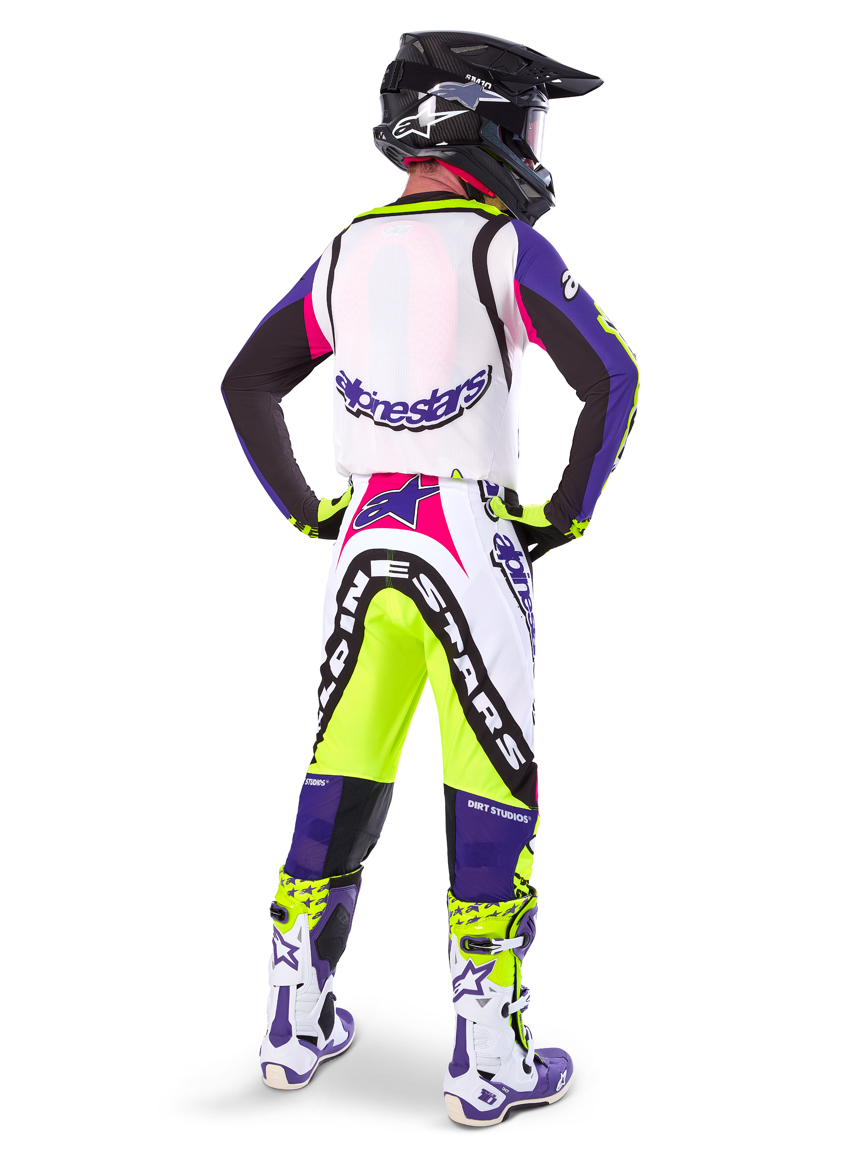 Motocross-Fahrer in Alpinestars Limited Edition Supertech Dirt Studios® Hose in Weiß, Lila und Gelb Fluo, Rückansicht mit passendem Trikot, Stiefeln und schwarzem Carbon-Helm, professionelle Offroad-Rennausrüstung mit markanten Grafiken und Neon-Akzenten.