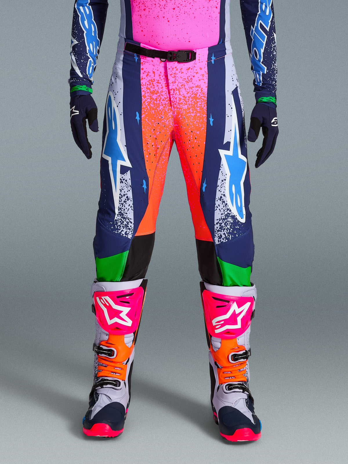 Alpinestars Limited Edition Supertech Records Hose, Motocross-Hose, Mehrfarbig mit lebendigen pink- und orangefarbenen Splash-Grafiken, dunkelblaue Einsätze mit blauen Stern-Logos und grünen Akzenten, kombiniert mit passendem Jersey sowie grau, pink und orangefarbenen Motocross-Stiefeln.