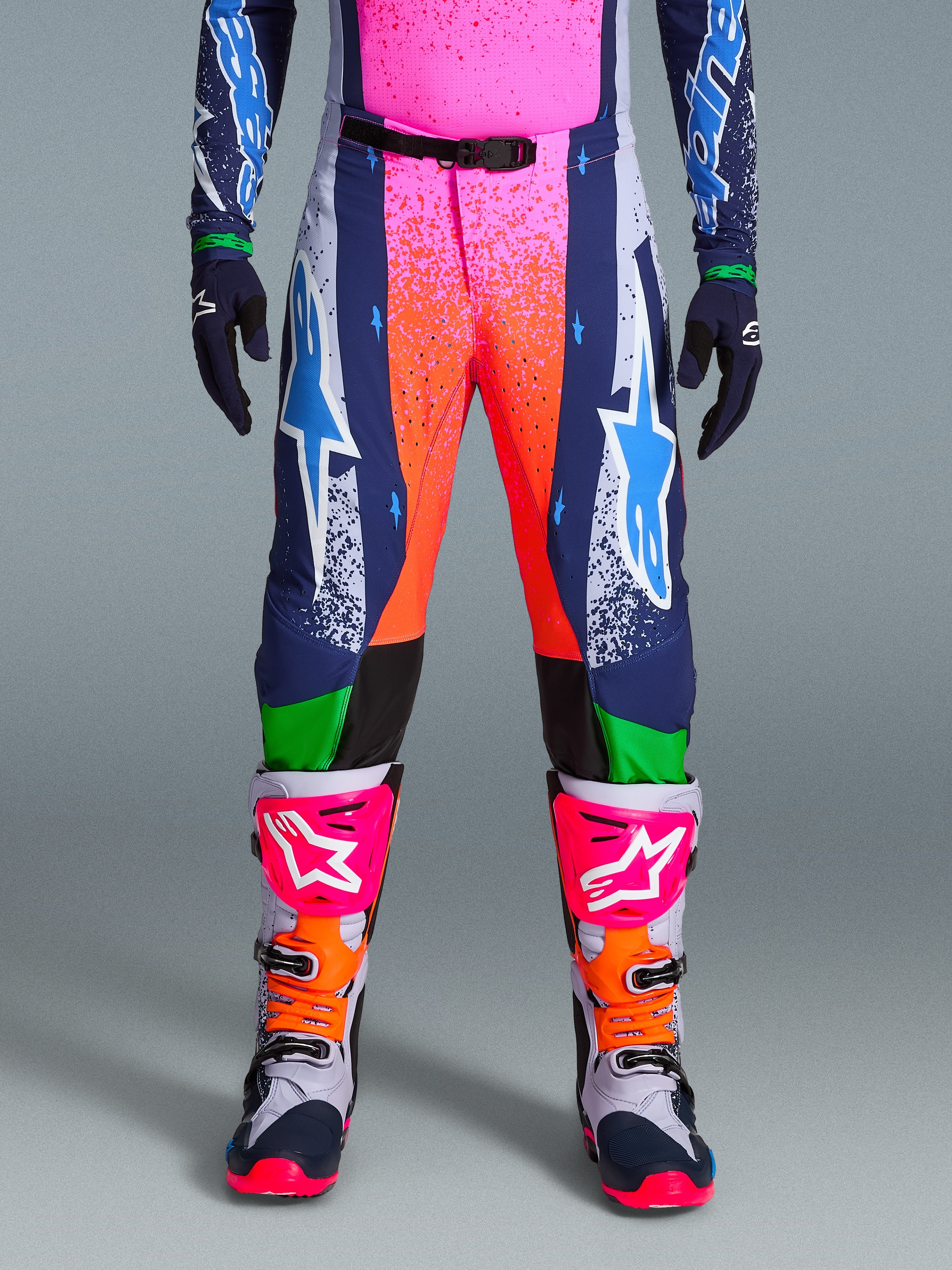 Alpinestars Limited Edition Supertech Records broek, motorcrossbroek, veelkleurig met levendige roze en oranje splash-graphics, donkerblauwe panelen met blauwe ster-logo's en groene accenten, gedragen met bijpassende jersey en grijze, roze en oranje motorcrosslaarzen.